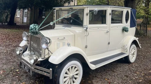 A cream vintage Imperial wedding car with white wall tyres, chrome grille, and teal bow decorations sits on a gravel driveway surrounded by trees and rustic buildings.
