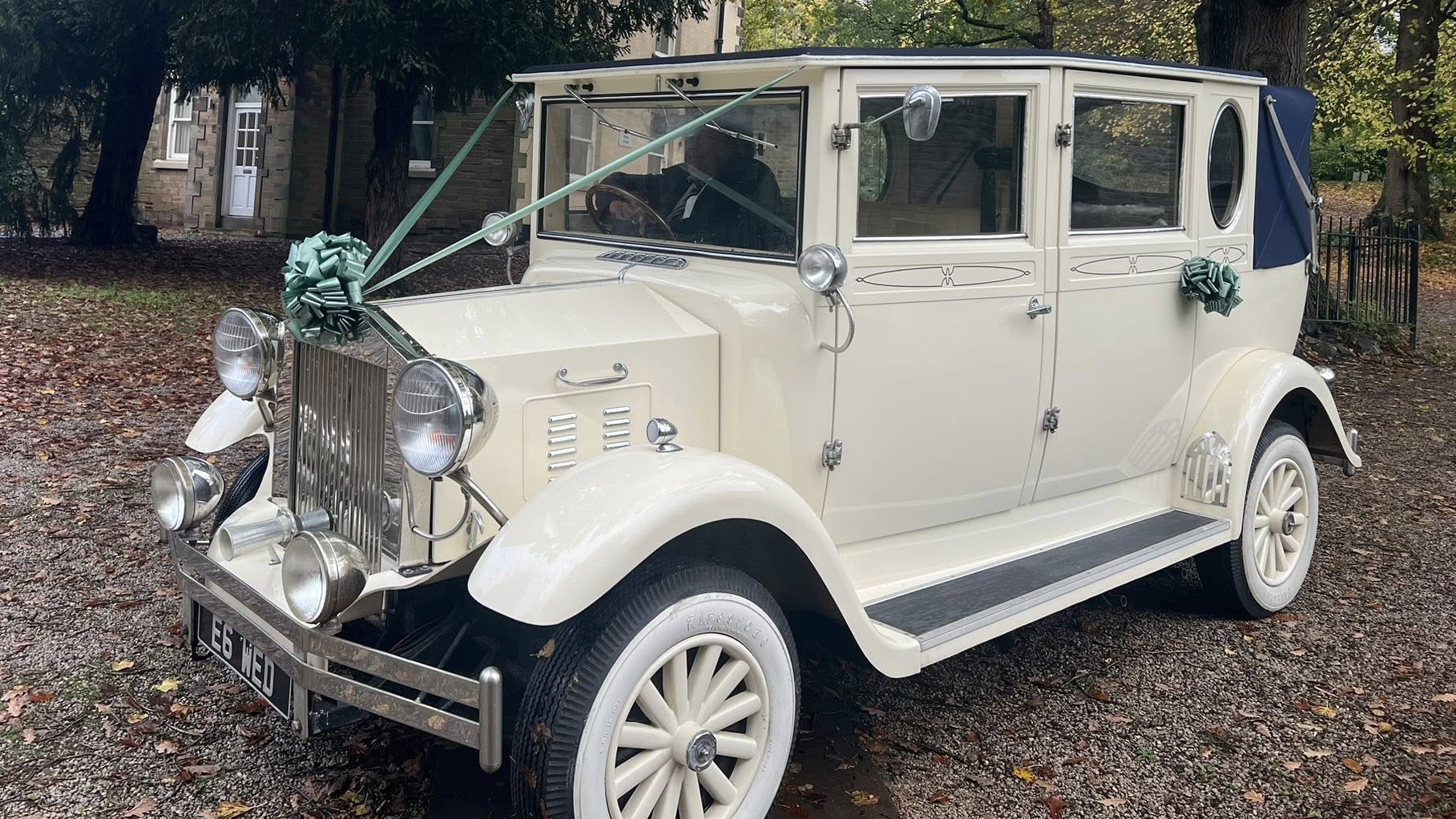 A cream vintage Imperial wedding car with white wall tyres, chrome grille, and teal bow decorations sits on a gravel driveway surrounded by trees and rustic buildings.