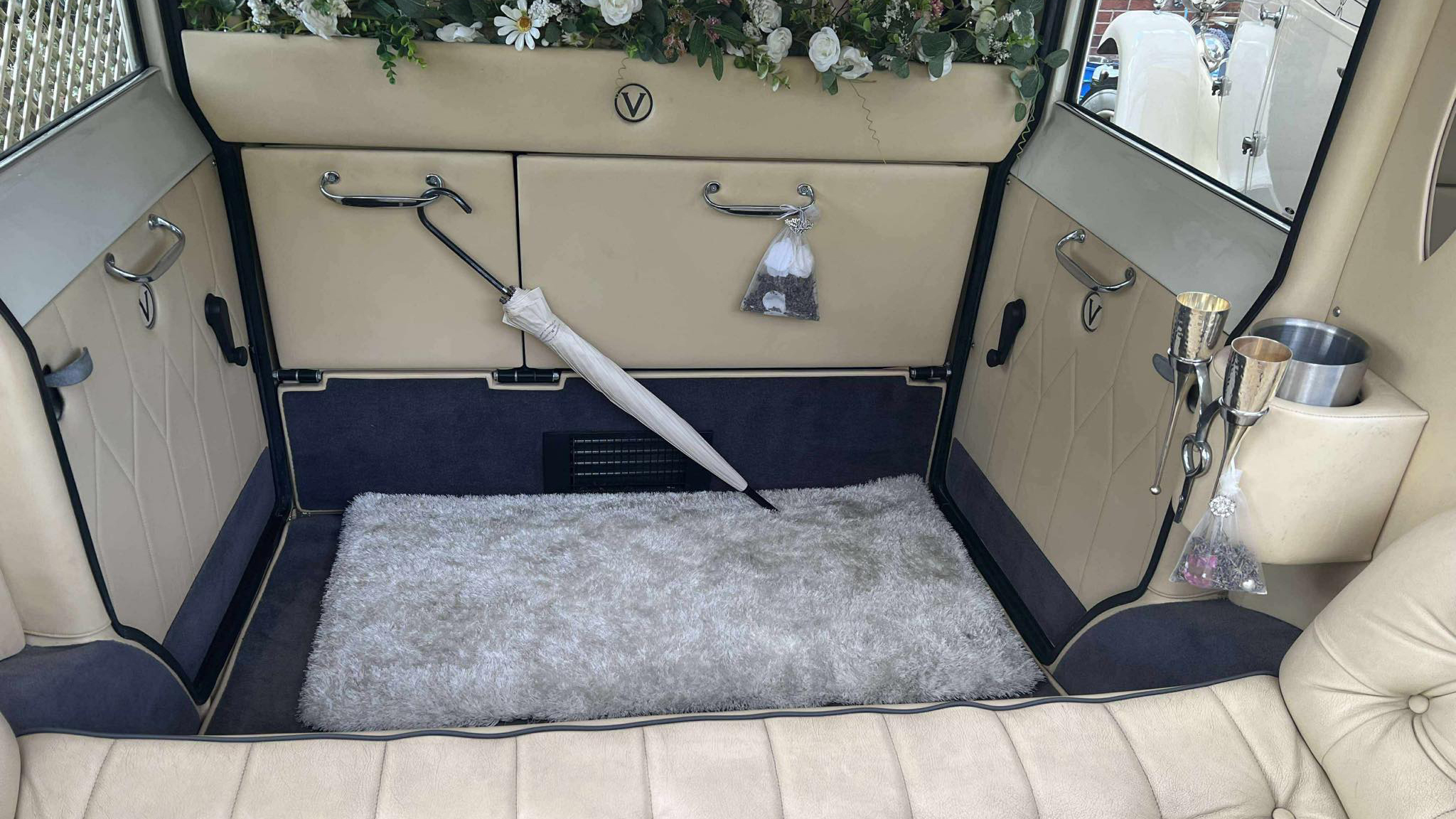 Interior view of the Viscount wedding car showing cream leather seating, button-back upholstery, polished trim and side floral decorations, with windows looking out onto greenery.