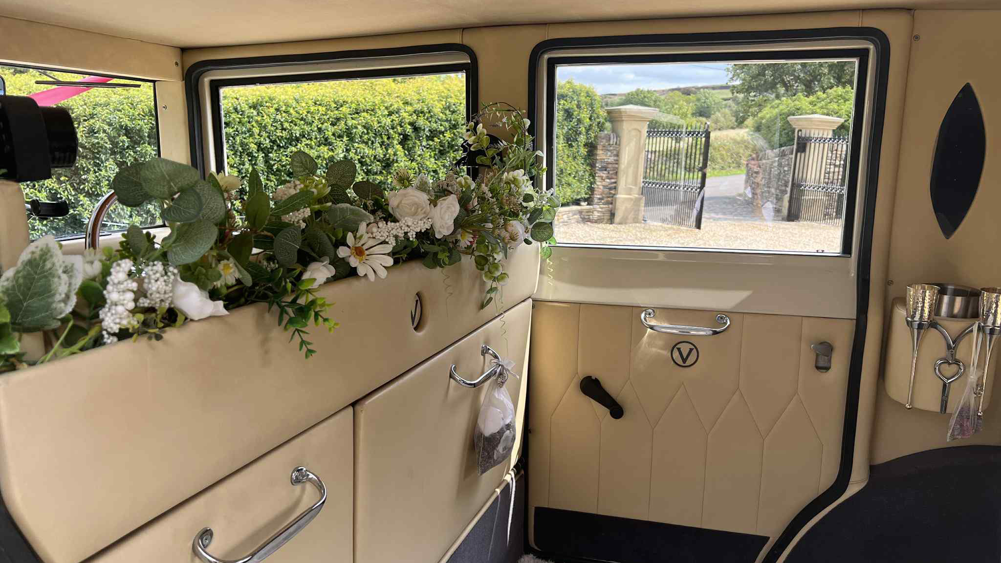 Close-up of cream leather seating inside a vintage wedding car with floral garlands, ivory panels and views of trees outside the window.