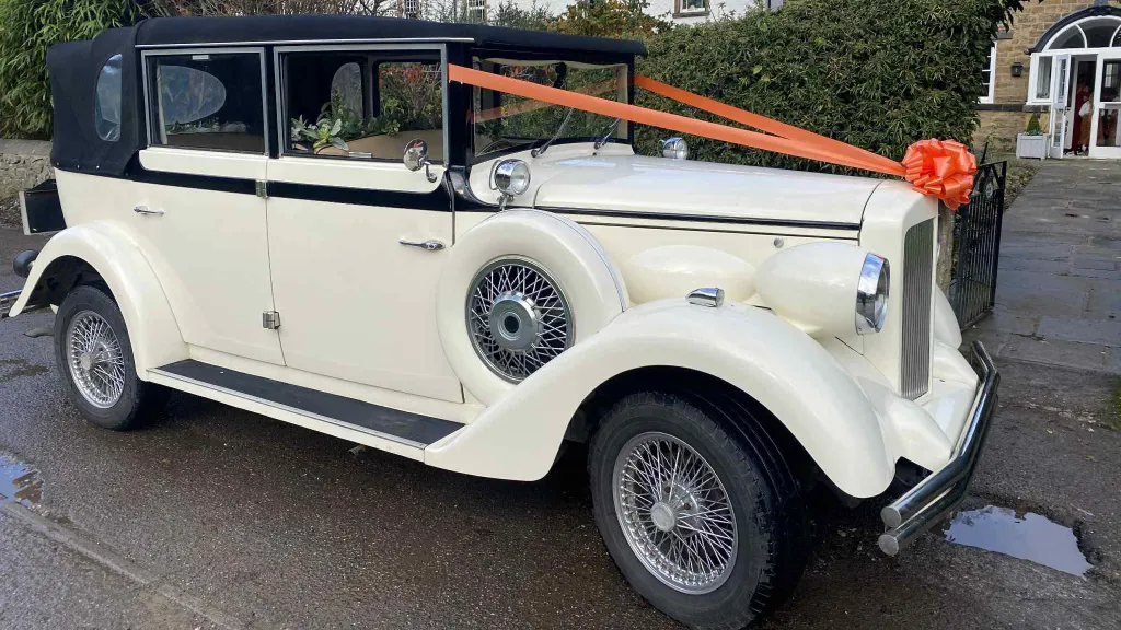 A cream and black vintage Regent wedding car photographed from the rear-side angle, decorated with a bright orange bow on the roof bar and featuring classic spare wheels on the side, parked on a wet driveway with garden seating in the background.