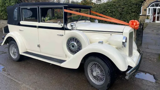 A cream and black vintage Regent wedding car photographed from the rear-side angle, decorated with a bright orange bow on the roof bar and featuring classic spare wheels on the side, parked on a wet driveway with garden seating in the background.