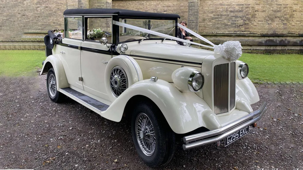 A cream and black vintage Regent wedding car viewed from the front-side angle, showing its distinctive chrome grille, landaulette roof, and spare wheel design, parked in front of a stone wall with a neatly cut lawn.