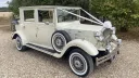 Front-side view of a cream Viscount vintage limousine decorated with white wedding ribbons and bows, featuring chrome headlamps, a side-mounted spare wheel and classic wire wheels, parked on gravel.