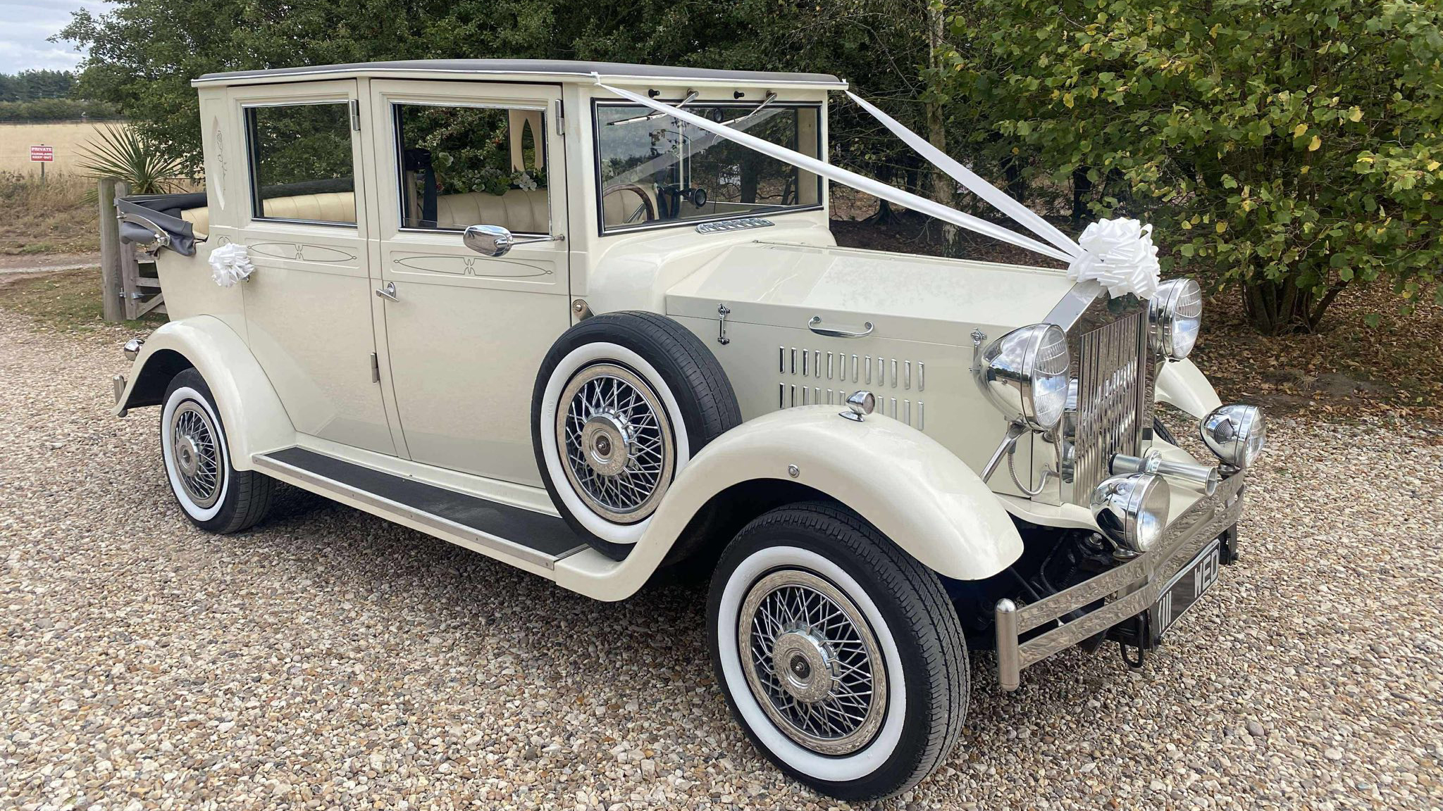Front-side view of a cream Viscount vintage limousine decorated with white wedding ribbons and bows, featuring chrome headlamps, a side-mounted spare wheel and classic wire wheels, parked on gravel.