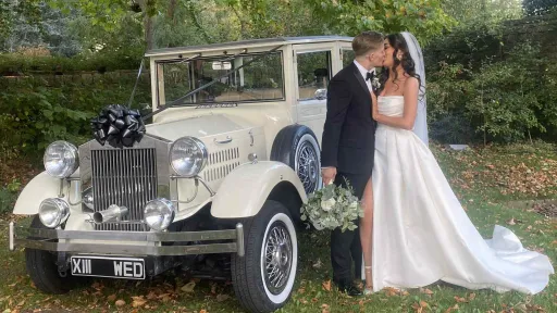 Bride and groom posing in front of a cream Viscount vintage wedding car with chrome grille and white ribbons, standing on a gravel driveway surrounded by trees.