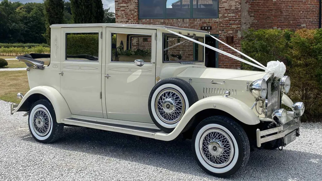 Side view of a cream Viscount vintage wedding car decorated with white ribbons and bows, highlighting the classic running boards and tinted windows, parked outdoors on a gravel surface.