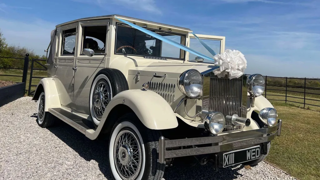 Cream Viscount vintage wedding car decorated with white bows and soft blue ribbons, featuring chrome detailing and spare wheels, parked beside a field under a cloudy sky.