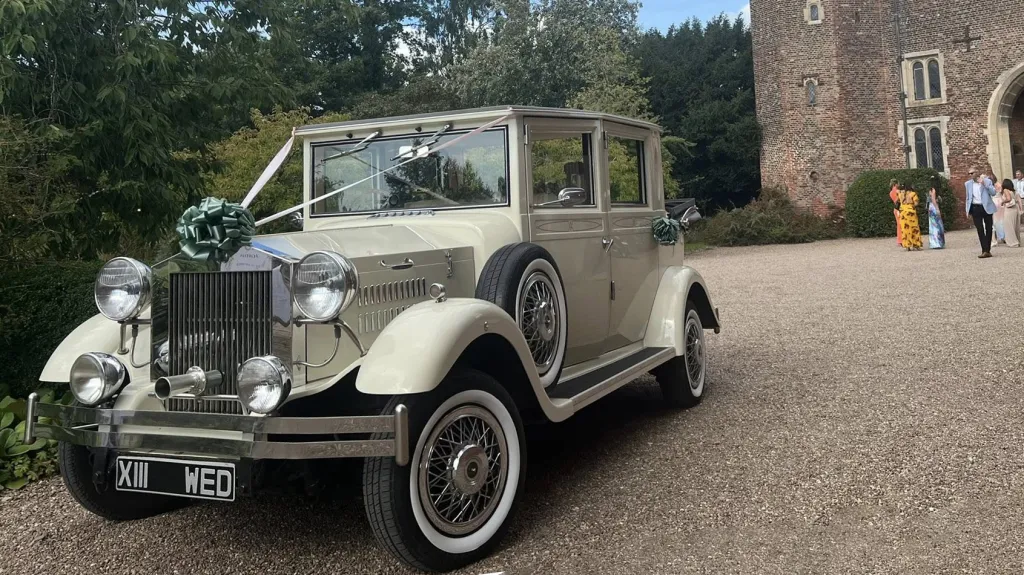 Cream and ivory Viscount limousine with vintage styling, decorated with white wedding ribbons and bows, parked on gravel with historic stone buildings in the background.