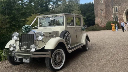 Cream and ivory Viscount limousine with vintage styling, decorated with white wedding ribbons and bows, parked on gravel with historic stone buildings in the background.