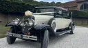Front-angled view of an ivory and black Bramwith wedding limousine decorated with ivory bows and ribbons. The vehicle shows its side-mounted spare tyre, tall chrome headlights, and retro body. A large country estate and greenery sit behind it.