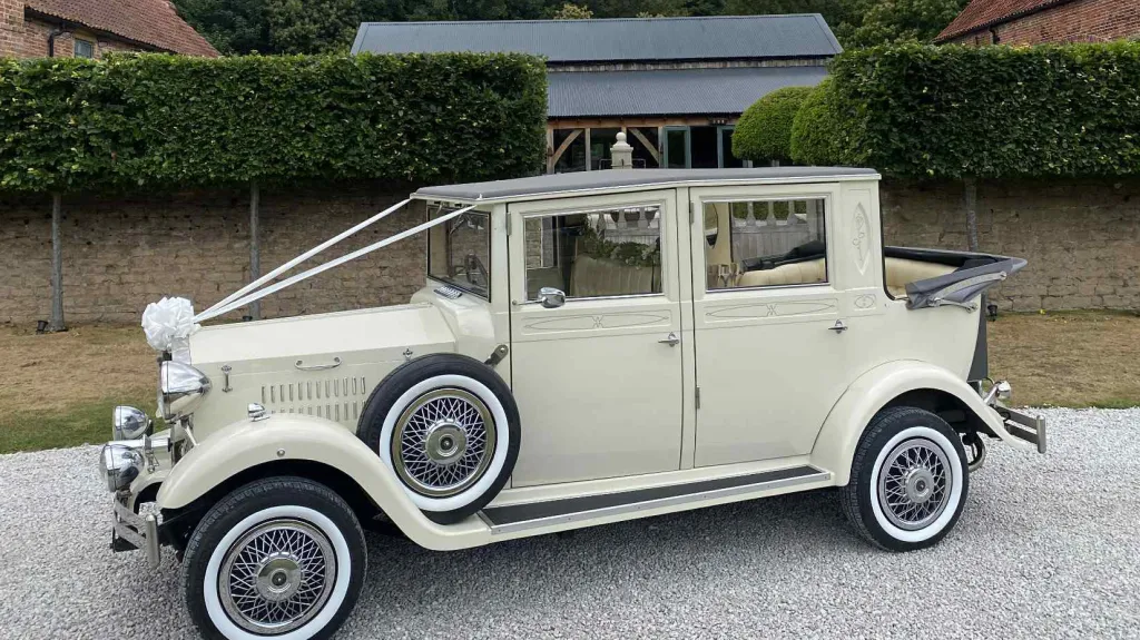 Left-side view of a cream vintage Imperial-style wedding car with white ribbons and bows, featuring chrome headlights and wire wheels, parked on gravel with hedges behind.