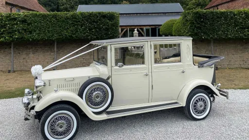 Left-side view of a cream vintage Imperial-style wedding car with white ribbons and bows, featuring chrome headlights and wire wheels, parked on gravel with hedges behind.