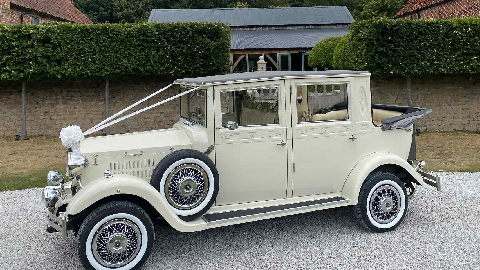 Left-side view of a cream vintage Imperial-style wedding car with white ribbons and bows, featuring chrome headlights and wire wheels, parked on gravel with hedges behind.