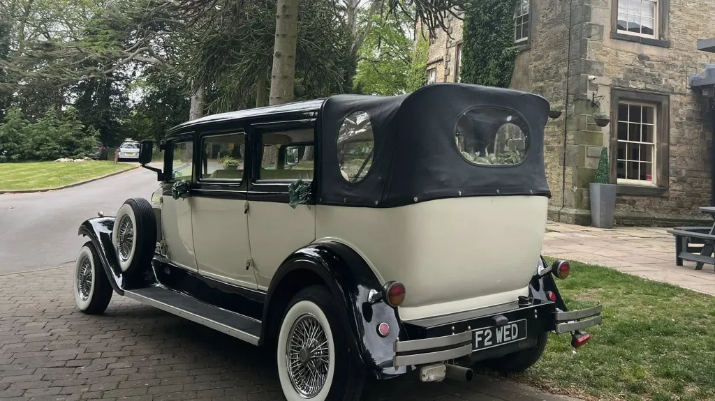 Rear view of an ivory and black vintage Bramwith limousine with a black roof and visible leather interior. The car is parked on a quiet road beside stone buildings and trees.