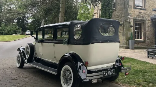 Rear view of an ivory and black vintage Bramwith limousine with a black roof and visible leather interior. The car is parked on a quiet road beside stone buildings and trees.