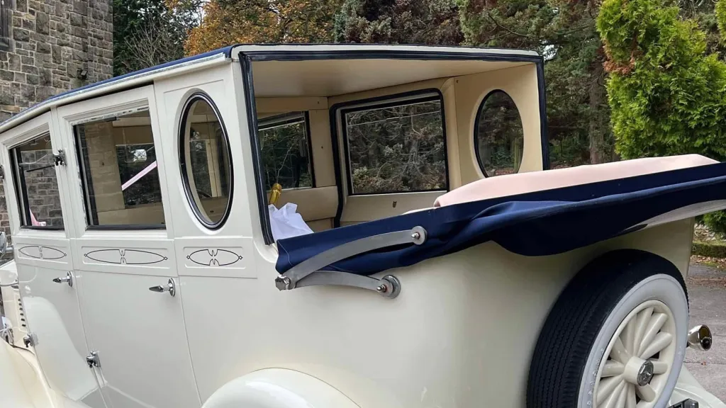 Rear view of a cream vintage Imperial wedding car with its blue landaulette-style roof partially folded back, showing cream leather interior seating. The vehicle is parked on a gravel driveway beside stone buildings and greenery.