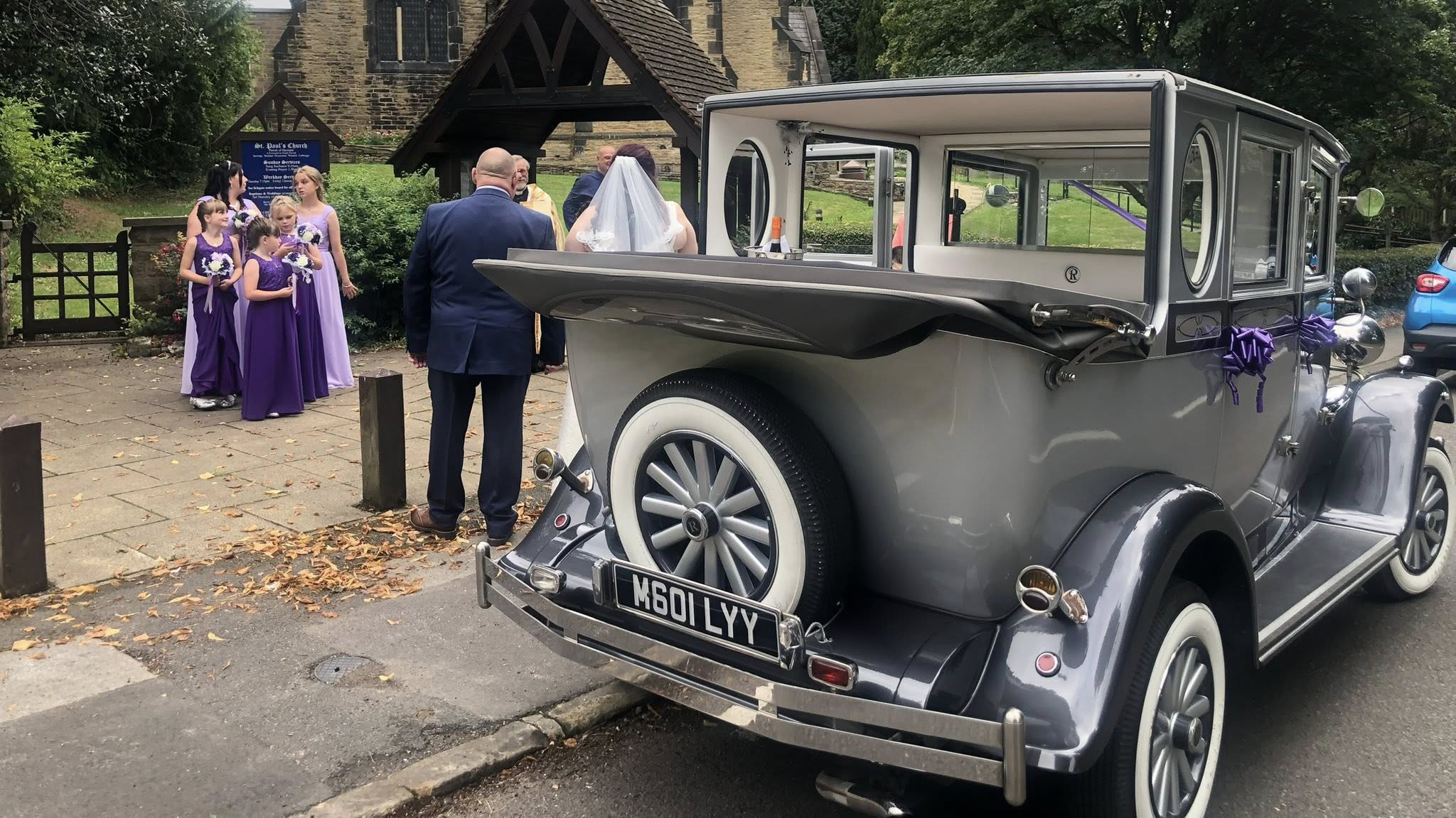 Rear view of a silver Imperial wedding car with the landaulette roof lowered. The spare wheel mounted on the back and white wall tyres are visible, with wedding guests dressed in colourful purple outfits standing in the background near a brick church entrance.