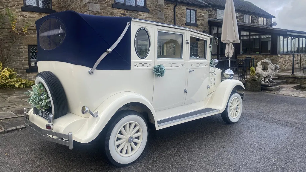Rear-side view of a cream Imperial vintage wedding car with its navy blue landaulette roof fully closed, showcasing its spare wheel, chrome bumper, and traditional design, parked outside a stone building with a lawn area.