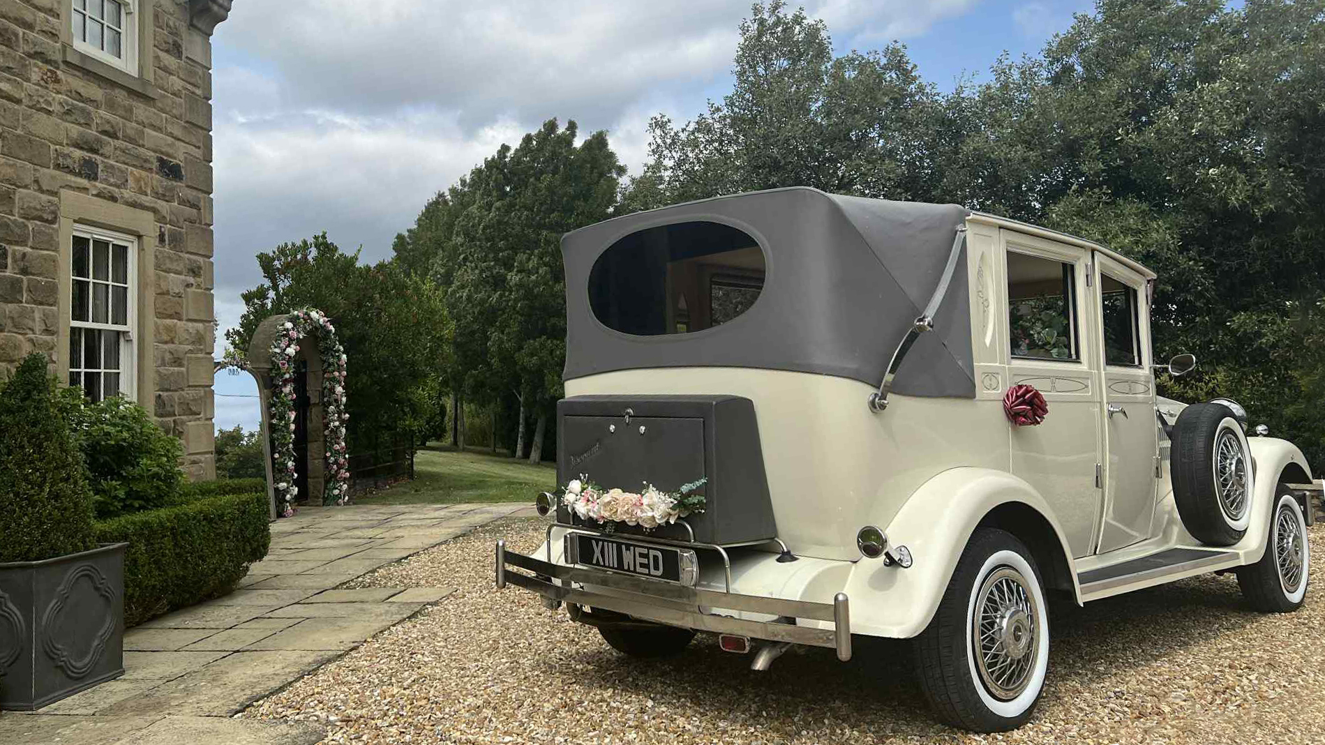 Rear view of a cream Viscount wedding car with a navy soft-top roof partially folded, classic oval rear windows and vintage chrome details, parked in front of a stone venue.