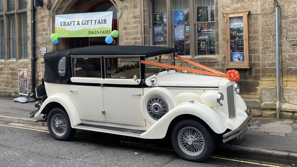 A right-side view of a cream and black vintage Regent wedding car decorated with an orange bow on the roof bar, featuring spare wheels, chrome bumpers and a classic design, parked in front of a building with signage.