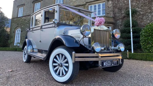 Front-view of a silver Imperial wedding car decorated with pink ribbons and bows across the bonnet and grille. The chrome headlights and vintage styling stand out, with a stone building and mature greenery in the background adding to the elegant scene.