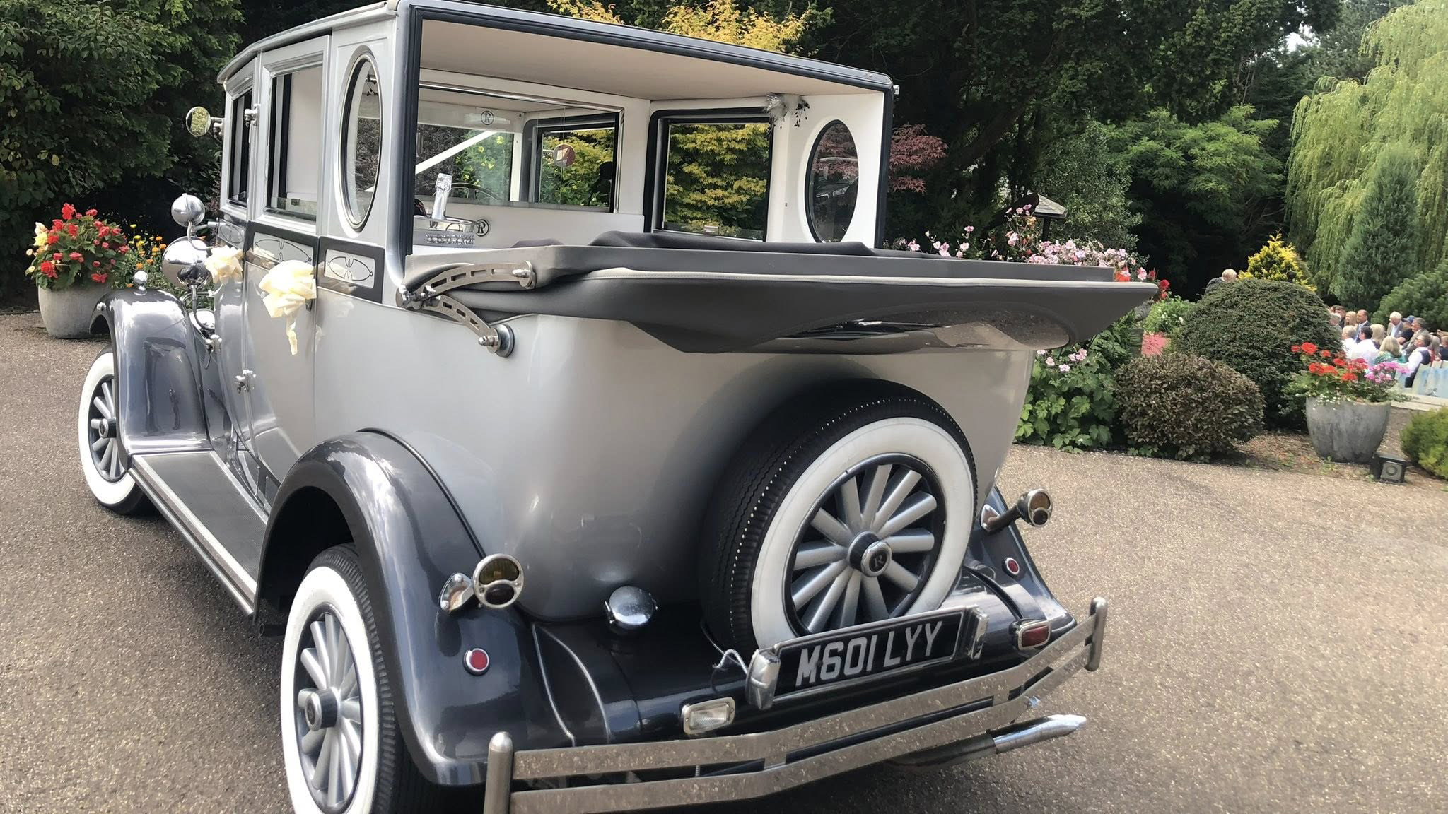 A silver and cream Imperial wedding car photographed from the front-left angle with the convertible rear roof folded down. The chrome headlights, twin spare wheels on the wings and white-spoked wheels give the car a classic 1930s appearance. The setting includes a garden with greenery and flowers.