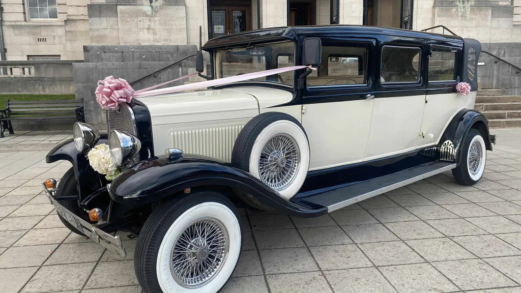 A classic ivory and black Bramwith wedding limousine decorated with pale pink bows is parked outside an old stone venue. The car’s side-mounted spare wheel, chrome grille, and whitewall tyres are clearly visible.