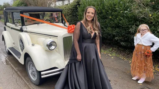 A bride wearing a black wedding dress standing beside a cream and black vintage Regent wedding car decorated with an orange bow, accompanied by a young bridesmaid in an orange dress, set on a tree-lined driveway with wet ground.