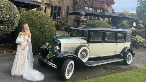 Side view of an ivory and black Bramwith limousine with a bride in a white dress standing beside it holding a colourful bouquet. The groom wears a dark suit. The car shows its spare wheel, chrome bumper, and cream interior. Background includes hedges, lawn, and a driveway.