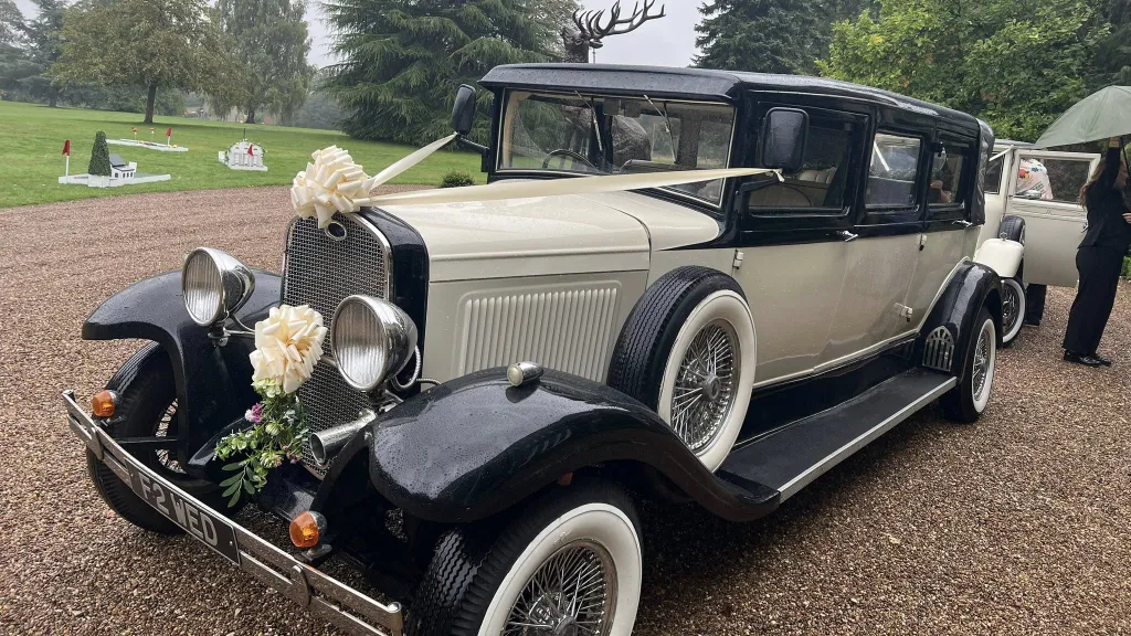 Front-side view of a black and ivory Bramwith limousine with side-mounted spare wheel, chrome lamps, and whitewall tyres. A wedding venue garden with grass and trees appears behind it.