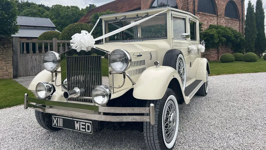 Cream vintage Viscount limousine decorated with a white bow and classic chrome grille, featuring side-mounted spare wheels, parked on gravel beside scenic landscaping.