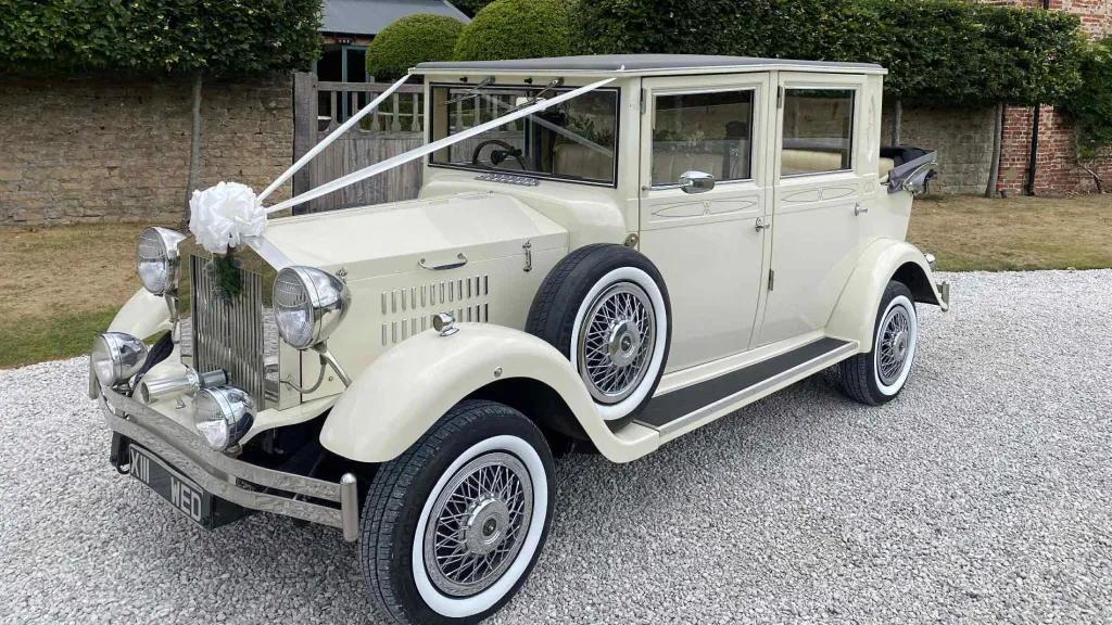Cream Viscount vintage wedding car with chrome headlights, white ribbons and full-length windows, parked outside a venue with greenery and trees.