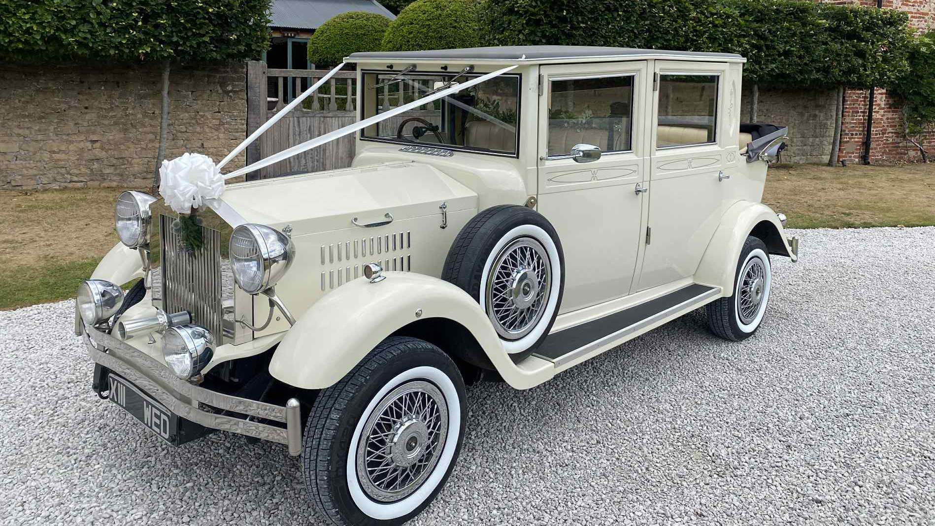 Cream Viscount vintage wedding car with chrome headlights, white ribbons and full-length windows, parked outside a venue with greenery and trees.