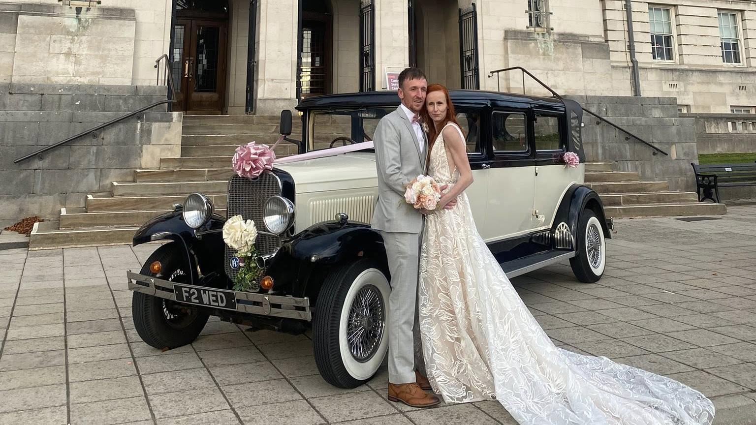 A bride in a long white gown and a groom in a dark suit stand beside a black and ivory Bramwith wedding limousine decorated with white ribbons. The car’s spare wheel and vintage chrome detailing are visible. The setting is a stone courtyard outside a grand building.