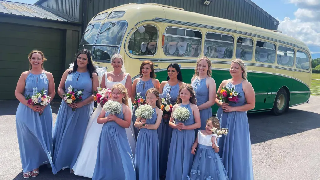 Bride and bridesmaids standing in front of a yellow and green vintage single-decker bus parked outside a courtyard building.