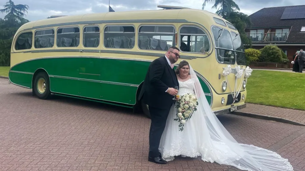 Yellow and green vintage single-decker bus shown from a side angle with the bride and groom embracing beside it on a stone driveway.