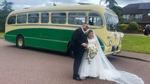Yellow and green vintage single-decker bus shown from a side angle with the bride and groom embracing beside it on a stone driveway.