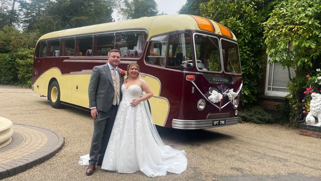 Burgundy and cream vintage single-decker bus photographed from the side, with a bride and groom standing beside it near a lakeside path.