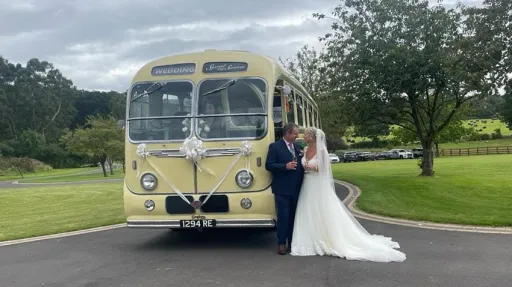 Yellow and green vintage single-decker bus photographed from the front angled view with the bride and groom posing beside it on a sunny day.