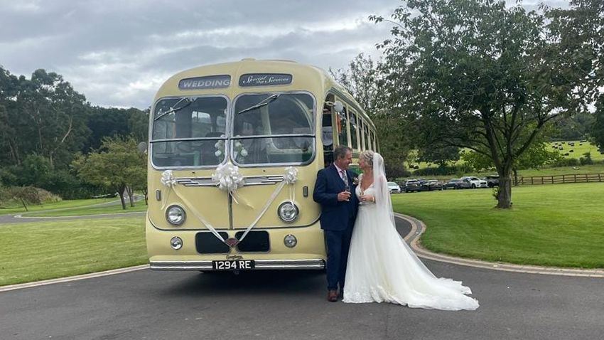 Yellow and green vintage single-decker bus photographed from the front angled view with the bride and groom posing beside it on a sunny day.
