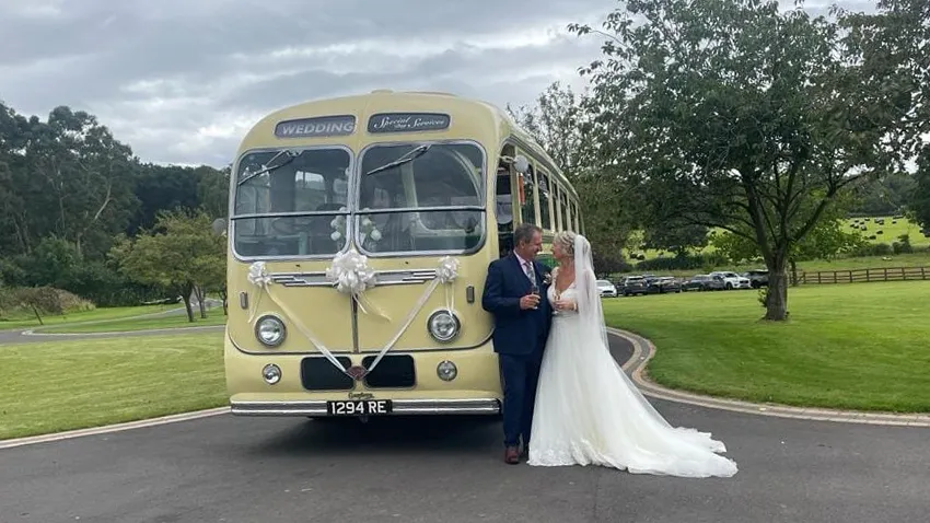 Yellow and green vintage single-decker bus photographed from the front angled view with the bride and groom posing beside it on a sunny day.