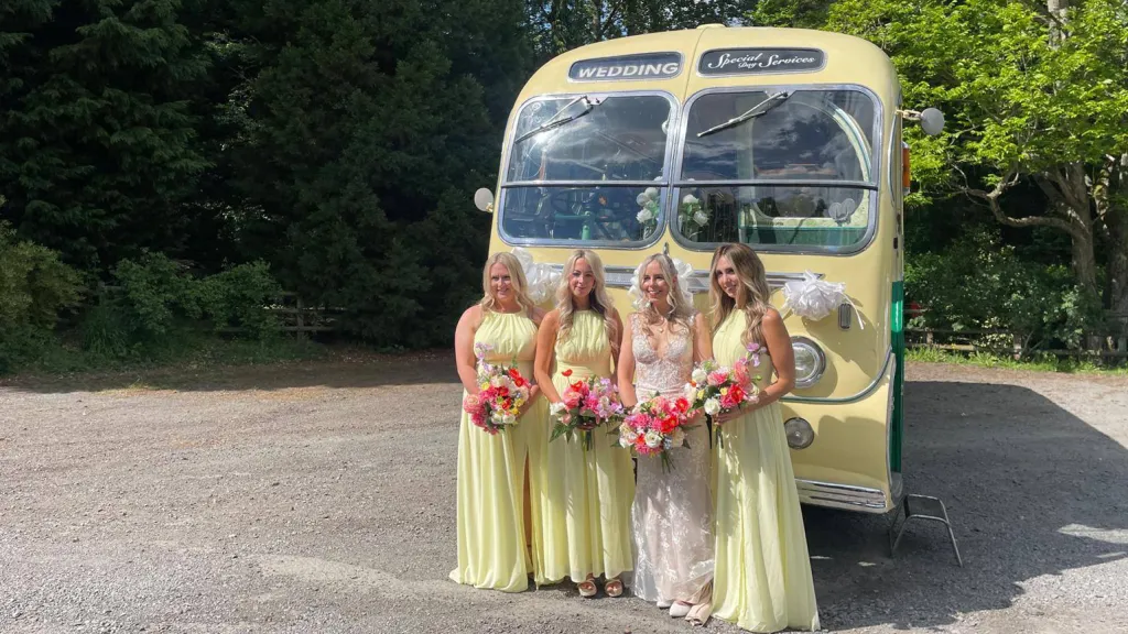 Bridesmaids in lemon-yellow dresses standing beside a yellow and green vintage single-decker bus in a shaded car park.
