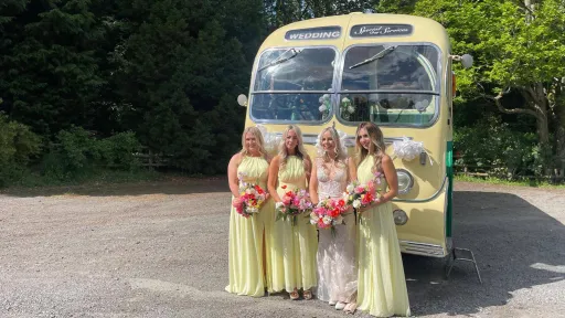 Bridesmaids in lemon-yellow dresses standing beside a yellow and green vintage single-decker bus in a shaded car park.