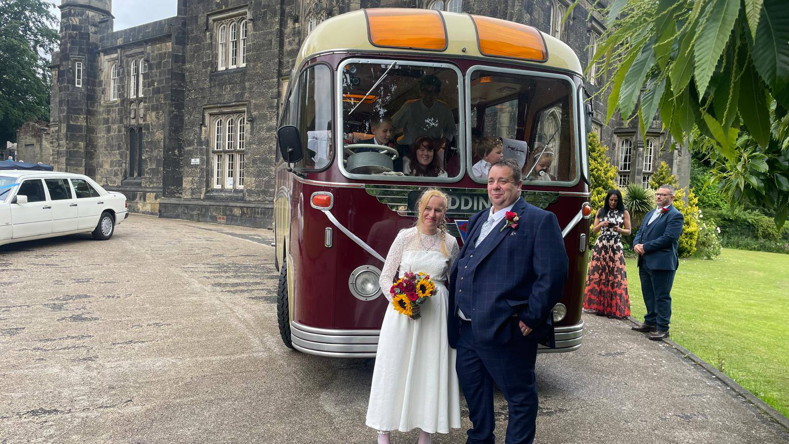 Front-facing burgundy and cream vintage single-decker bus parked outside a historic stone building, with a bride and groom in front.