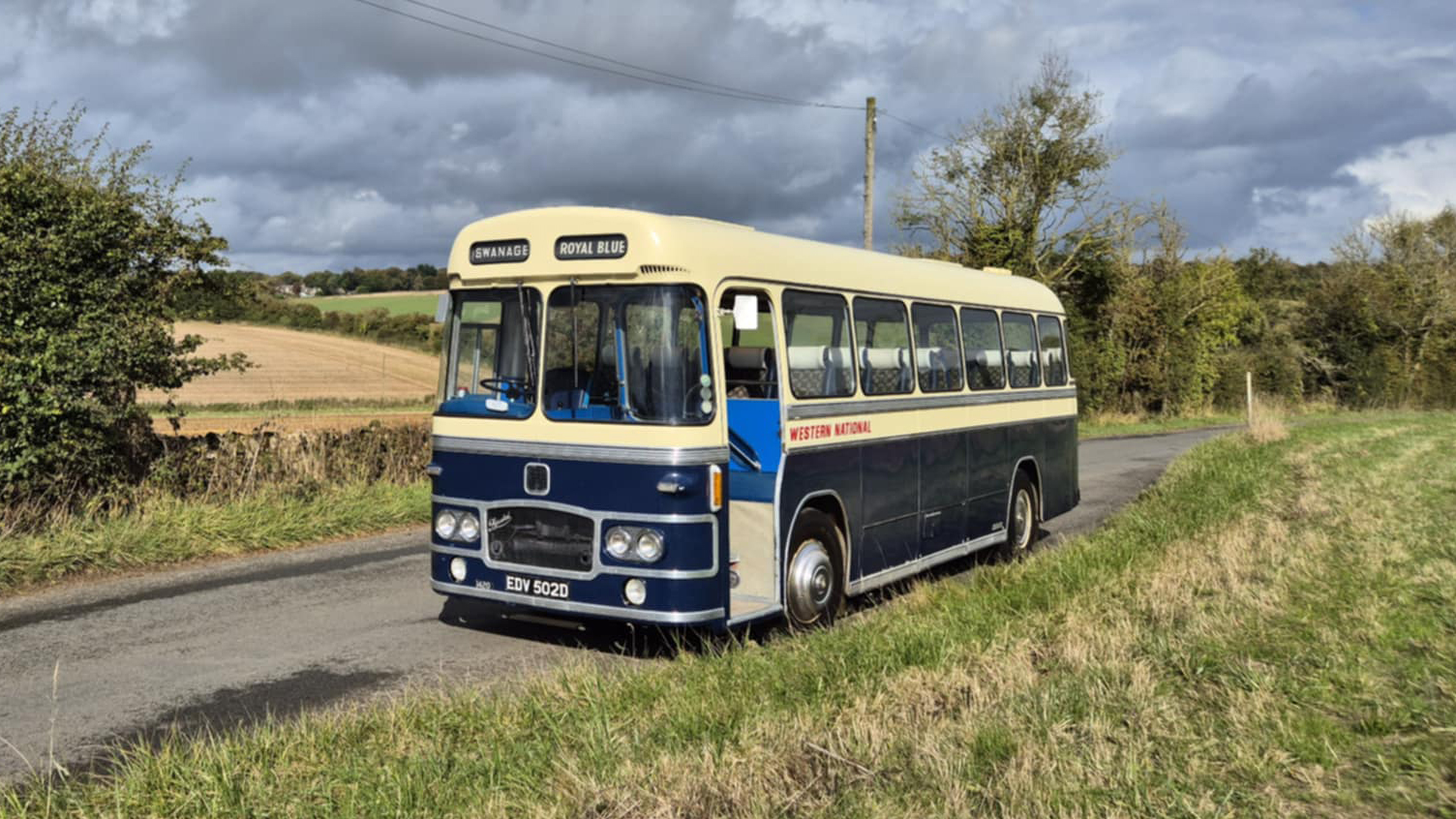 Blue and ivory vintage single-decker bus photographed from a front angled view, parked on a rural road with open fields and trees behind.