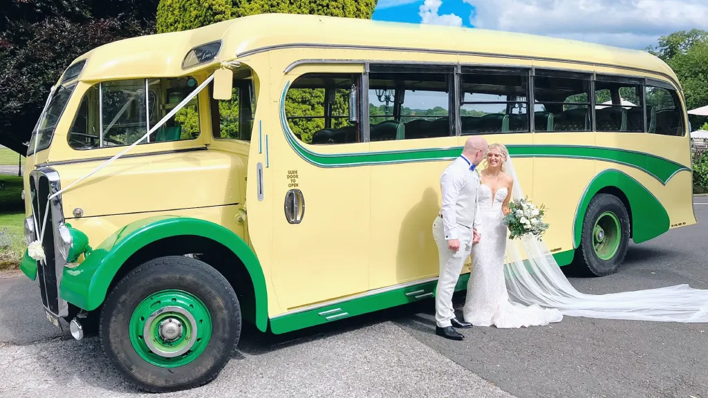 Yellow and green vintage single-decker bus shown from a front angled view, parked beside a bride near a stone courtyard.