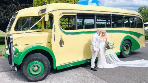Yellow and green vintage single-decker bus shown from a front angled view, parked beside a bride near a stone courtyard.
