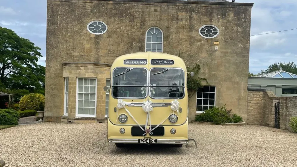 Front-facing view of a yellow and green vintage single-decker bus parked on a gravel courtyard with a manor house behind.