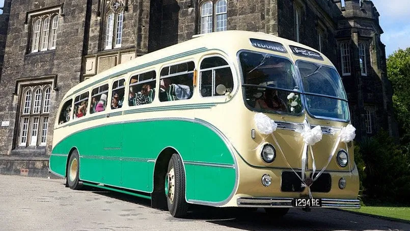 Green and yellow vintage single-decker bus shown from a side-front angle outside a large stone venue on a sunny day.